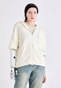 Kremfarget hettegenser med glidelås foran, som har store lommer og korte ermer, sammen med lyseblå stripete jeans.