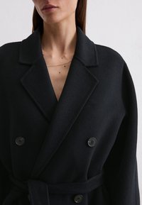 Black, double-breasted wool coat with a large collar, featuring two black buttons and a wrapped tie belt. Soft, textured fabric detail.
