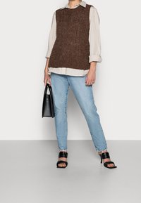 Brauner gestrickter ärmelloser Pullover über einem hellbeigen Hemd, kombiniert mit hellblauen Jeans und schwarzen Sandalen mit Absatz, hält eine schwarze Handtasche.