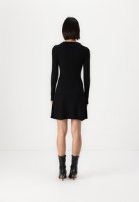 Robe noire à manches longues en ribbed avec un ourlet évasé, fabriquée à partir d'un matériau extensible. Portée avec des bottes noires à talons montant jusqu'aux genoux.