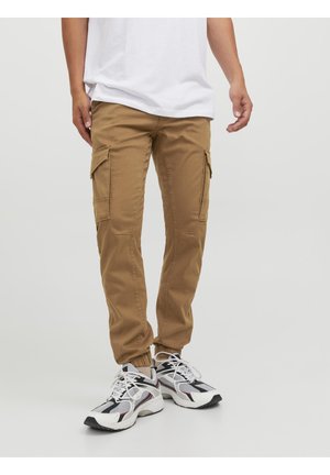 Mężczyzna w beżowych spodniach cargo z elastycznymi mankietami i białych sportowych sneakersach, stojący na jednolitym szarym tle.