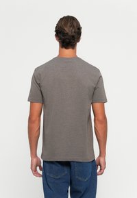Mann med kort brunt hår iført en ensfarget grå t-skjorte med korte ermer og blå jeans, sett fra baksiden mot en hvit bakgrunn.