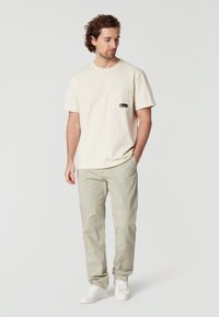 Crème-kleurige katoenen t-shirt met een borstzak en een klein zwart label, gecombineerd met lichtgrijze broek en witte sneakers.