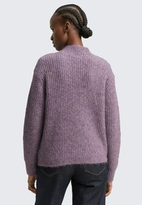 Fioletowy sweter dziergany z teksturowanym wzorem, żebrowanym dekoltem i lekko skróconym tyłem, w połączeniu z ciemnoniebieskimi dżinsami.