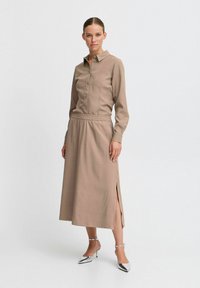 Vestido marrón de botón con cuello, cinturilla elástica, aberturas laterales, mangas largas y textura suave. Combinado con tacones plateados.