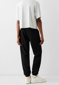 Svarta sweatpants med avslappnad passform, elastisk midja och sidoficka, kombinerat med en kortärmad ljusgrå topp och beige sneakers.