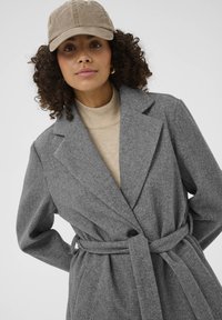 Manteau trench en mélange de laine gris avec une taille ceinturée, des revers en pointe et des manches longues. Porté avec un col roulé beige et une casquette beige.