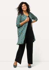Kimono lungo a stampa floreale in turchese, con punti multicolori. Indossato sopra un completo nero, con maniche a tre quarti e una vestibilità rilassata.