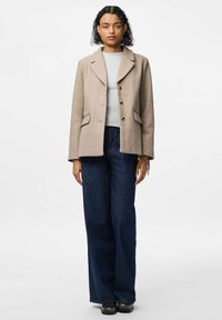 Heled pruun, villasegistusega ülerüü, millel on nurgelised kraed, nööbiefekt ja kaks klapiga taskut. Kantud halli ribakujulise ülakeha ja tumedate teksade peal.