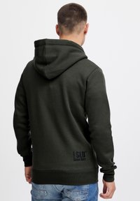 Felpa verde scuro realizzata in tessuto texture, con un grande cappuccio, polsini a costine e un logo con la scritta "Dal 1993" ricamata in basso.