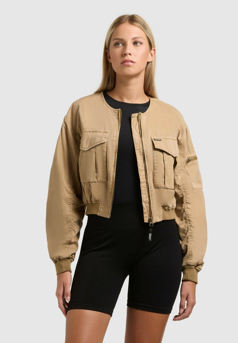Femme portant une veste bomber beige courte sur un short noir ajusté et un haut, debout devant un fond gris clair uni.