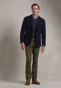 Ung man som står, klädd i marinblå kavaj över grå sweater och blå skjorta, olivgröna byxor och bruna finskor mot en neutral bakgrund.