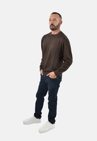 Maglione marrone con trama, jeans blu scuro e sneakers bianche. Il modello è in piedi con le mani nelle tasche, con un'espressione neutra.