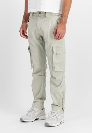 Mann trägt hellbeige Cargohosen mit mehreren Taschen und weiße Sportschuhe, steht vor einem schlichten weißen Hintergrund.