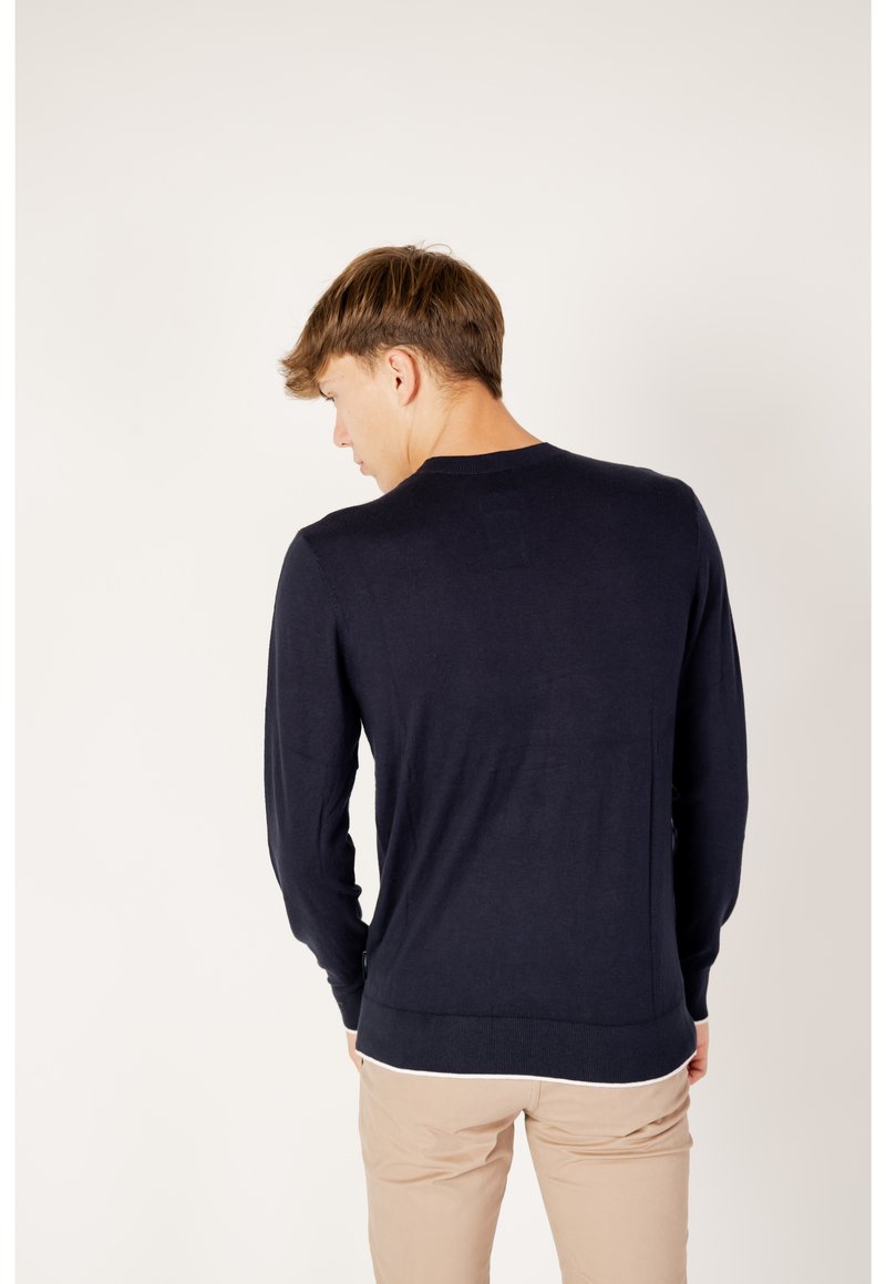 Junger Mann, der von hinten zu sehen ist, trägt einen navyblauen Langarmpullover und beige Hosen vor einem schlichten weißen Hintergrund.
