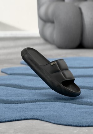 Zwarte slidesandaal met een getextureerde zool, rustend op een blauw tapijt, naast een vaag grijze gecushioneerde fauteuil in een minimalistische binnenruimte.