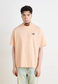 T-shirt oversized em cor pêssego, feito de algodão, com um logo preto da Adidas no lado esquerdo do peito. Mangas curtas, decote arredondado e ajuste descontraído.