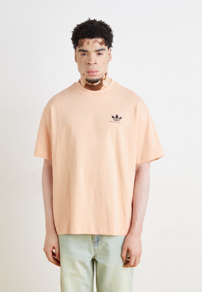 T-shirt oversized em cor pêssego, feito de algodão, com um logo preto da Adidas no lado esquerdo do peito. Mangas curtas, decote arredondado e ajuste descontraído.