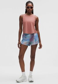 Träningsoutfit med en rosa ärmlös topp och iriserande blå shorts; modell som bär solglasögon och vita sneakers.