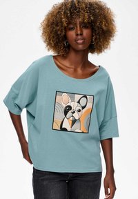 Korte mouwen, lichtblauw T-shirt met een grote geborduurde afbeelding van een Franse bulldog, met geometrische patronen in tinten zwart, oranje en crème.