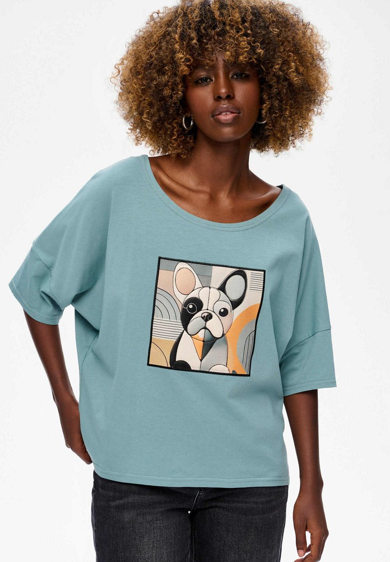 Korte mouwen, lichtblauw T-shirt met een grote geborduurde afbeelding van een Franse bulldog, met geometrische patronen in tinten zwart, oranje en crème.