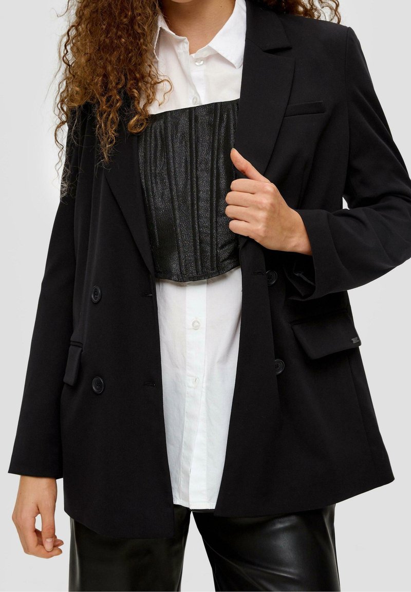 Femme portant une chemise blanche boutonnée, un corset noir texturé, un blazer noir oversize et un pantalon en cuir noir, tenant le blazer ouvert.