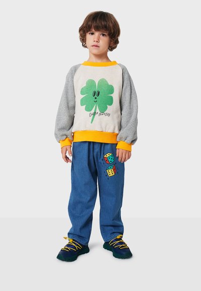 Sudadera gris con gráfico de trébol verde; cuello y puños amarillos. Pantalones anchos azules con diseño de dados coloridos; zapatos negros y amarillos.