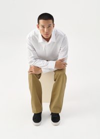 Hvit skjorte med lyse striper, khaki bukser og svarte joggesko. Modell sitter på en kube, med hendene sammenfoldet rundt knærne.