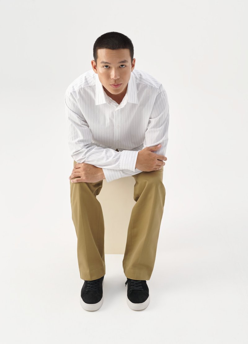 Hvit skjorte med lyse striper, khaki bukser og svarte joggesko. Modell sitter på en kube, med hendene sammenfoldet rundt knærne.