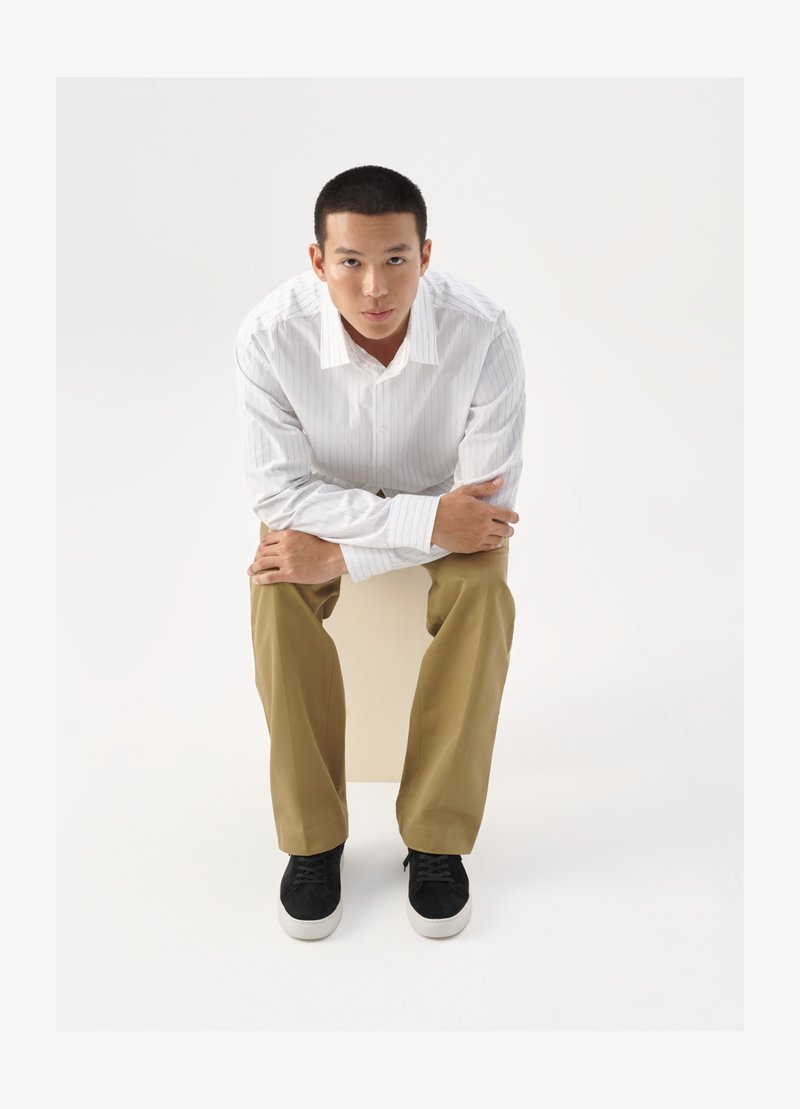 Hvit skjorte med lyse striper, khaki bukser og svarte joggesko. Modell sitter på en kube, med hendene sammenfoldet rundt knærne.