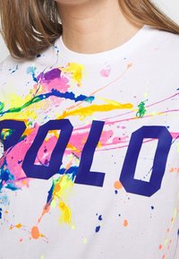 Bílá bavlněná tričko s velkým tmavě modrým logem "POLO". Tričko zobrazuje živé skvrny růžové, žluté a modré barvy.