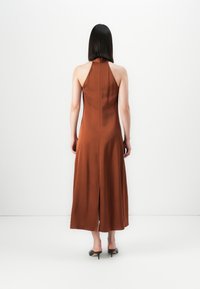 Kvinne som står med ryggen til kameraet, iført en ermeløs rust-brun halterneck midi-kjole og svarte kitten heels mot en hvit bakgrunn.