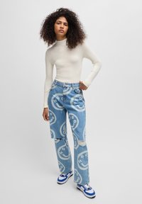 Lys kremfarget ribbestrikket langermet topp parret med blå slitte jeans med store hvite smilefjesmønstre, båret med blå sneakers.