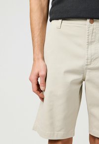 Beige bomuldsshorts med lige snit, der har en lille logoaccent og sidelommer. Teksturen er glat, og stoffet virker holdbart.