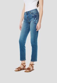 Blauwe spijkerbroek met bloemrijk borduurwerk op de linkerzak, gecombineerd met bruine sandalen met bandjes, tegen een effen grijze achtergrond.