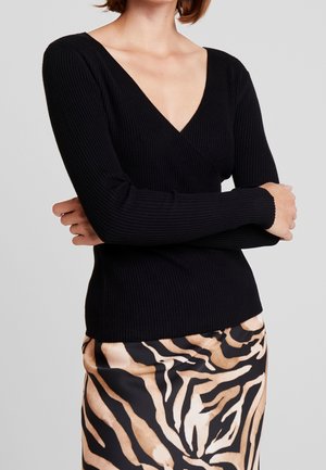 Kobieta ubrana w czarny prążkowany top z długim rękawem i dekoltem w serek oraz satynową spódnicę w beżowo-czarne wzory zebra, ze skrzyżowanymi ramionami.