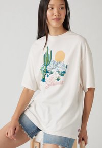 Camiseta blanca de gran tamaño con una colorida escena del desierto que incluye cactus, montañas y un gráfico del sol. Material de algodón suave, diseño de cuello redondo.