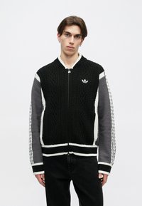 Mladý muž s čiernym, šedým a bielym pleteným zipsom na bunde s logom Adidas, stojí na svetlom pozadí.