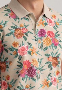 Květinová polo košile s béžovým pozadím obsahuje živé vyšívané vzory multicolor květin a zelených listů; klasický design límce.