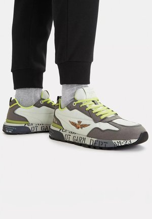 Sneakers bianche e grigie con lacci e dettagli giallo neon, indossate con calzini grigi e pantaloni neri, con logo e dettagli stampati sulla suola.