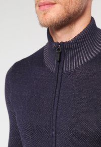 Granatowy sweter z zamkiem błyskawicznym, posiadający teksturowane dzianinowe body oraz prążkowany kołnierz, z zapięciem na zamek i subtelnymi różnicami w strukturze materiału.