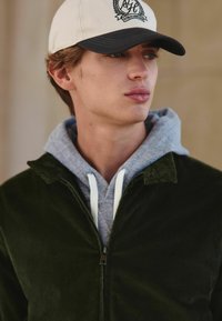 Groene corduroyjas over een grijze hoodie met witte trekkoorden, gecombineerd met een beige pet met een zwart logo en een gebogen klep.