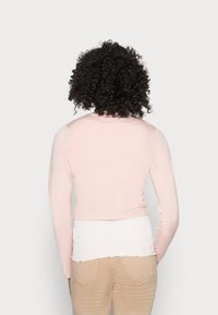 Femme aux cheveux bouclés portant un haut à manches longues rose pâle sur une chemise blanche et un pantalon beige, de dos sur un fond uni.