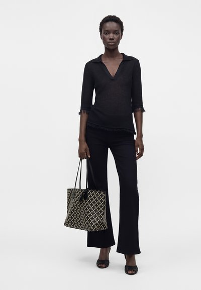 Femme portant un haut noir à col en V avec manches et ourlet à franges, pantalon noir, sandales noires, tenant un sac fourre-tout tissé à motif.