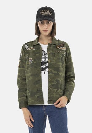 Femme portant une veste camouflage avec des patches, un t-shirt graphique blanc, un jean bleu et une casquette noire Von Dutch, debout devant un fond uni.
