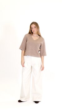 Žena stojaca, oblečená v béžovom svetri s výstrihom do V a s rukávmi dĺžky do lakťov, bielych širokých nohaviciach a tmavých topánkach na bielom pozadí.