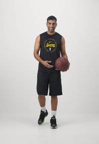 Camisola preta sem mangas com o logotipo amarelo dos "Lakers", calções pretos e sapatos pretos com detalhes verdes. A segurar uma bola de basquetebol, a sorrir.