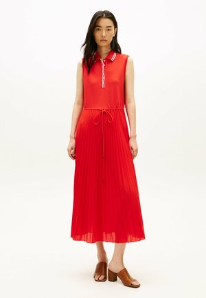 Kvinde iført rød poloskjole uden ærmer med plisseret nederdel og bindebånd i taljen, kombineret med brune sandaler med åben tå og blokhæl, stående mod hvid baggrund.