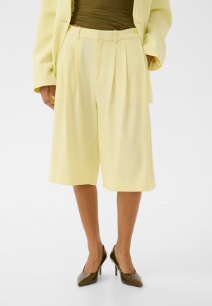 Femme portant un short ample jaune pâle au niveau du genou, un blazer assorti, un haut vert olive et des chaussures à talons hauts marron foncé.