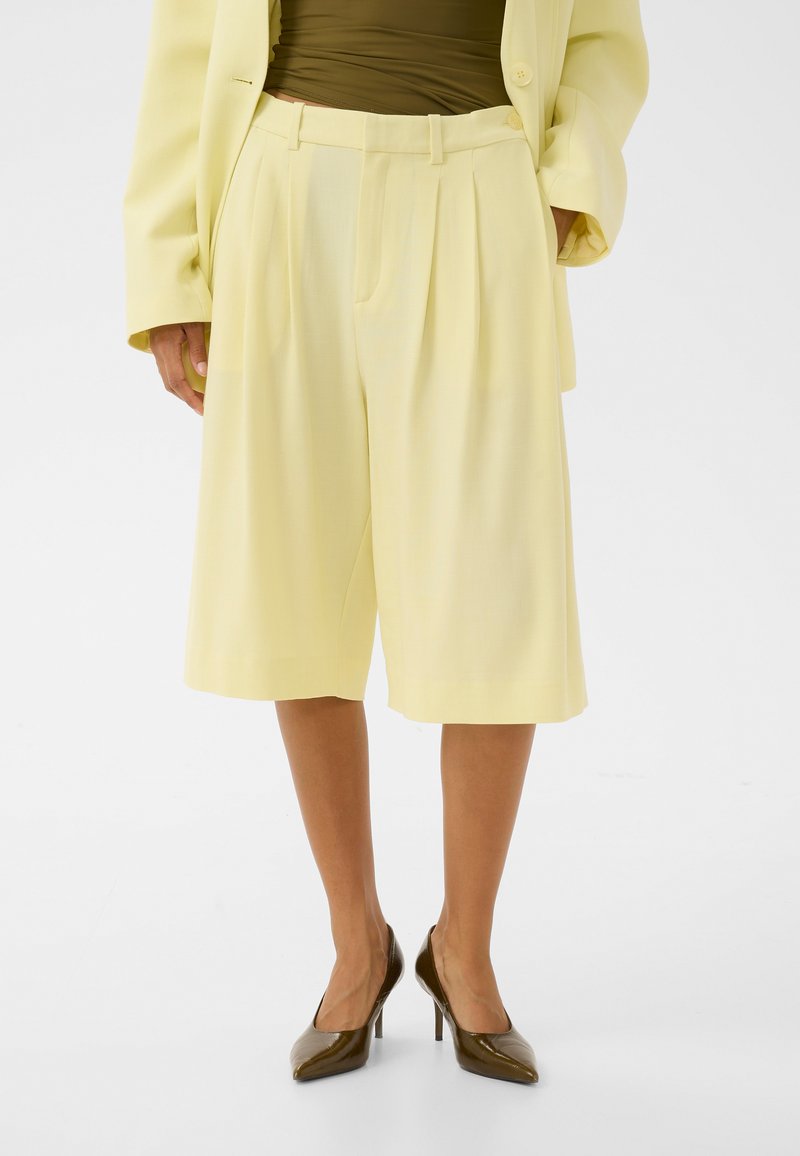 Femme portant un short ample jaune pâle au niveau du genou, un blazer assorti, un haut vert olive et des chaussures à talons hauts marron foncé.